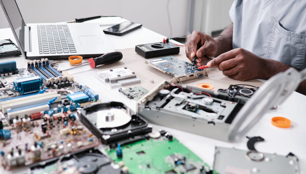 Hands of young African man repairing circuit board with soldering iron while sitting by desk in front of laptop by pressmaster